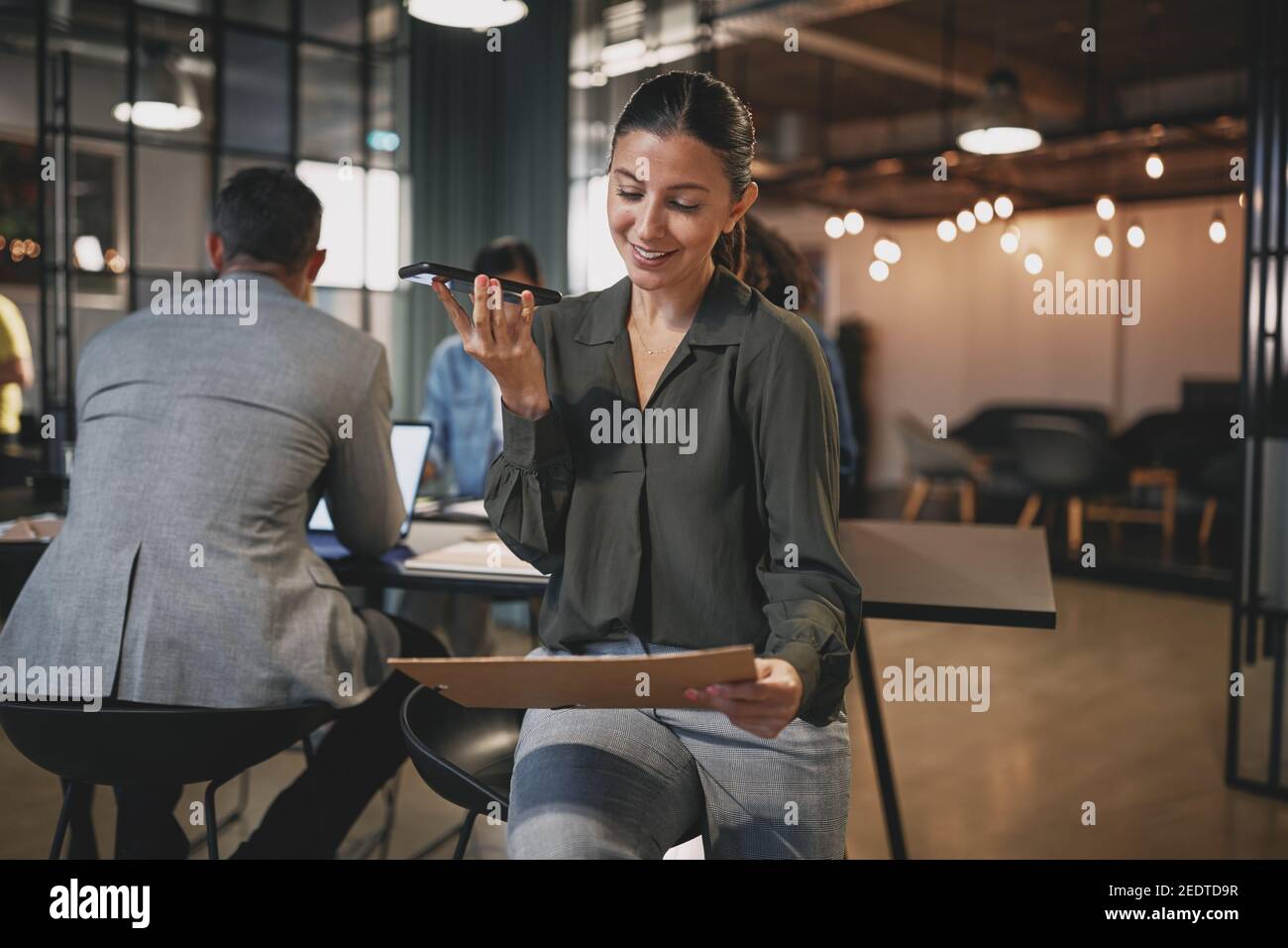 Smiling businesswoman reading paperwork and talking on speakerphone to a client with colleagues sitting in the background Stock Photo