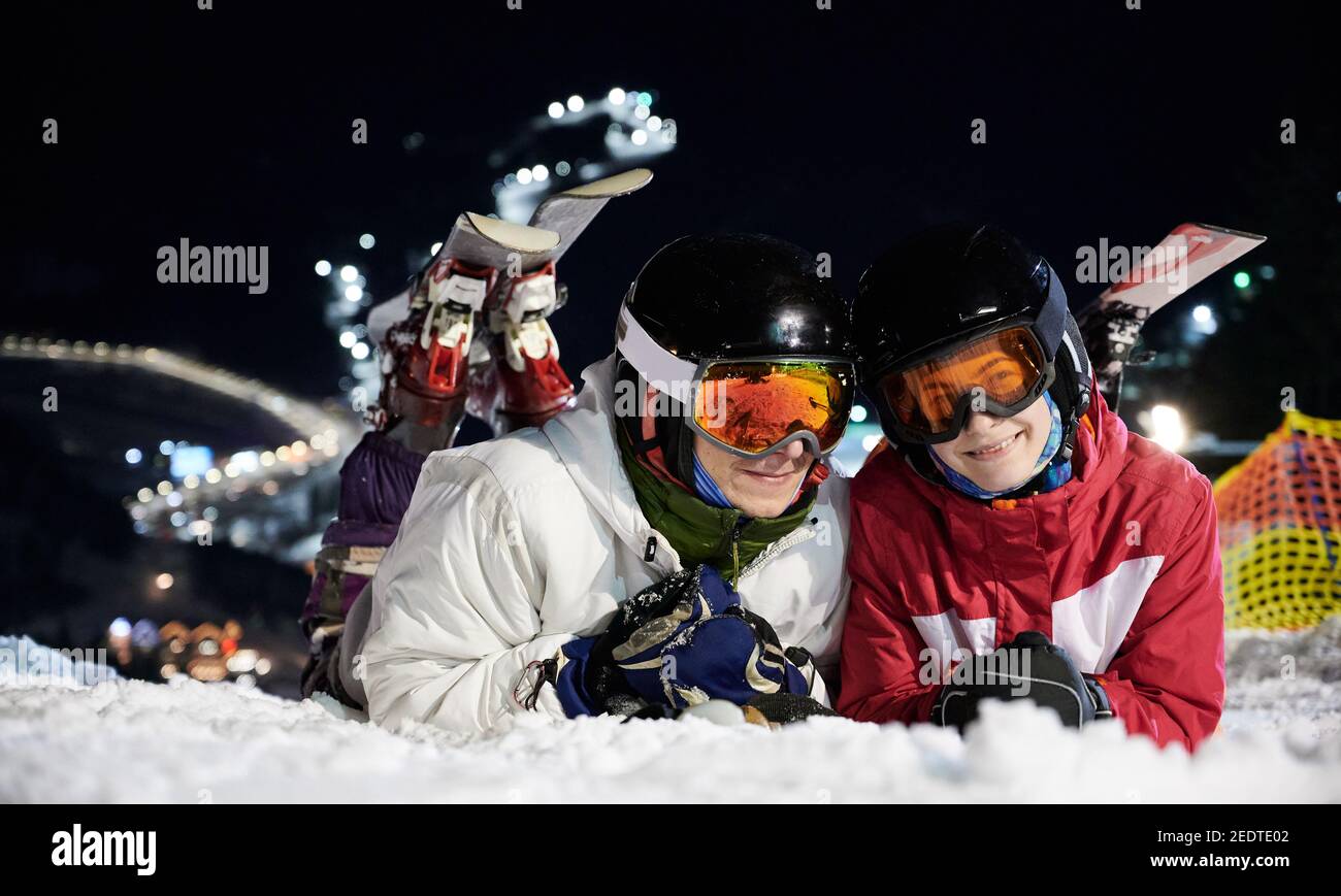 Couple of skiers lying on snowy slope under illuminated chairlift at night, resting after active time spending on ski pistes. Close-up portrait of two cheerful happy people on snow Stock Photo