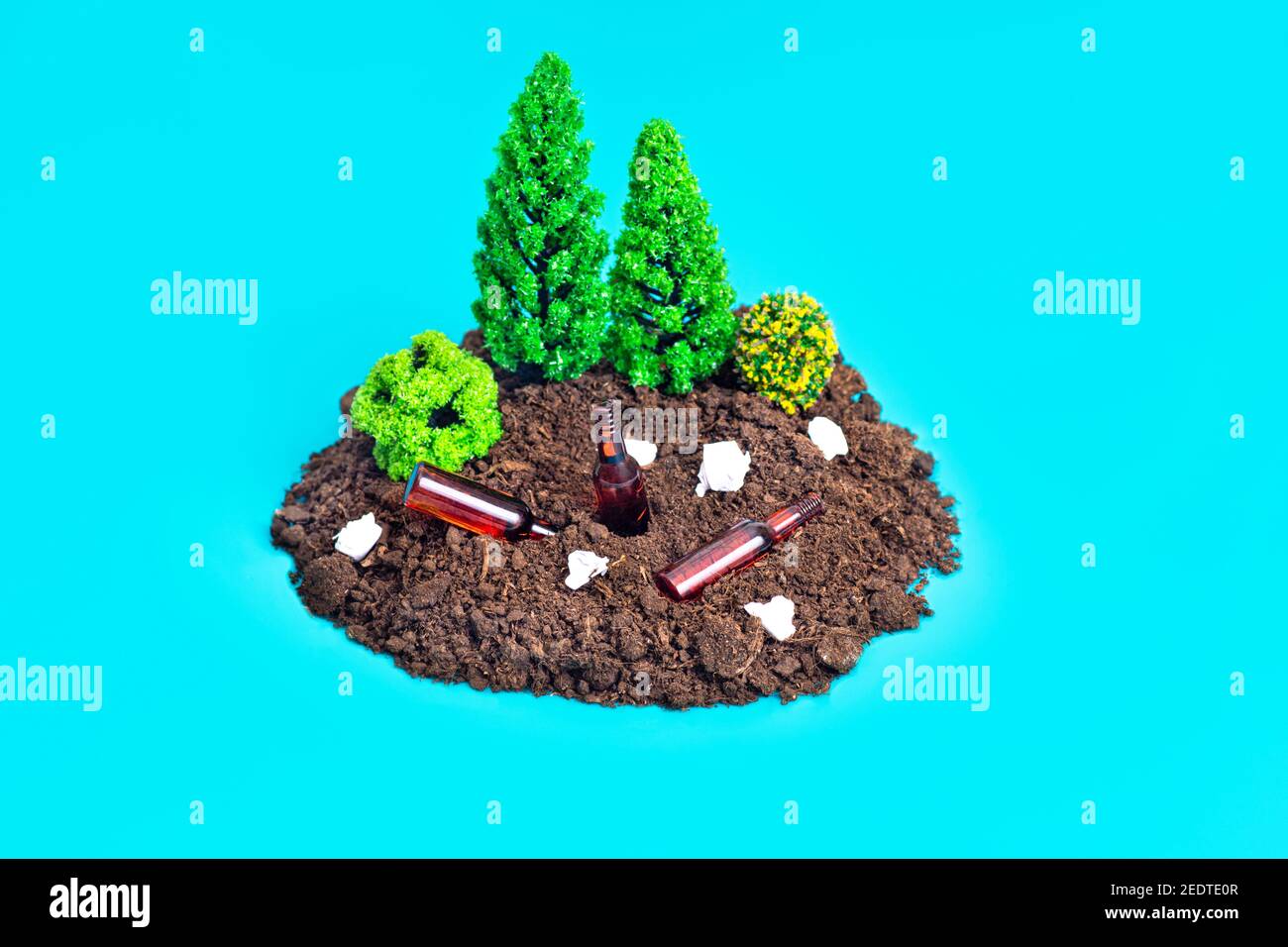 Miniature toy forest composition on a blue background with paper rubbish and tiny beer bottles lying around. Environmental impact concept. Stock Photo