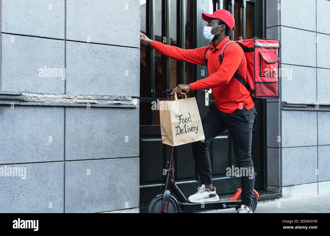 African rider man delivering meal to customers with electric scooter while wearing face mask during corona virus outbreak Stock Photo