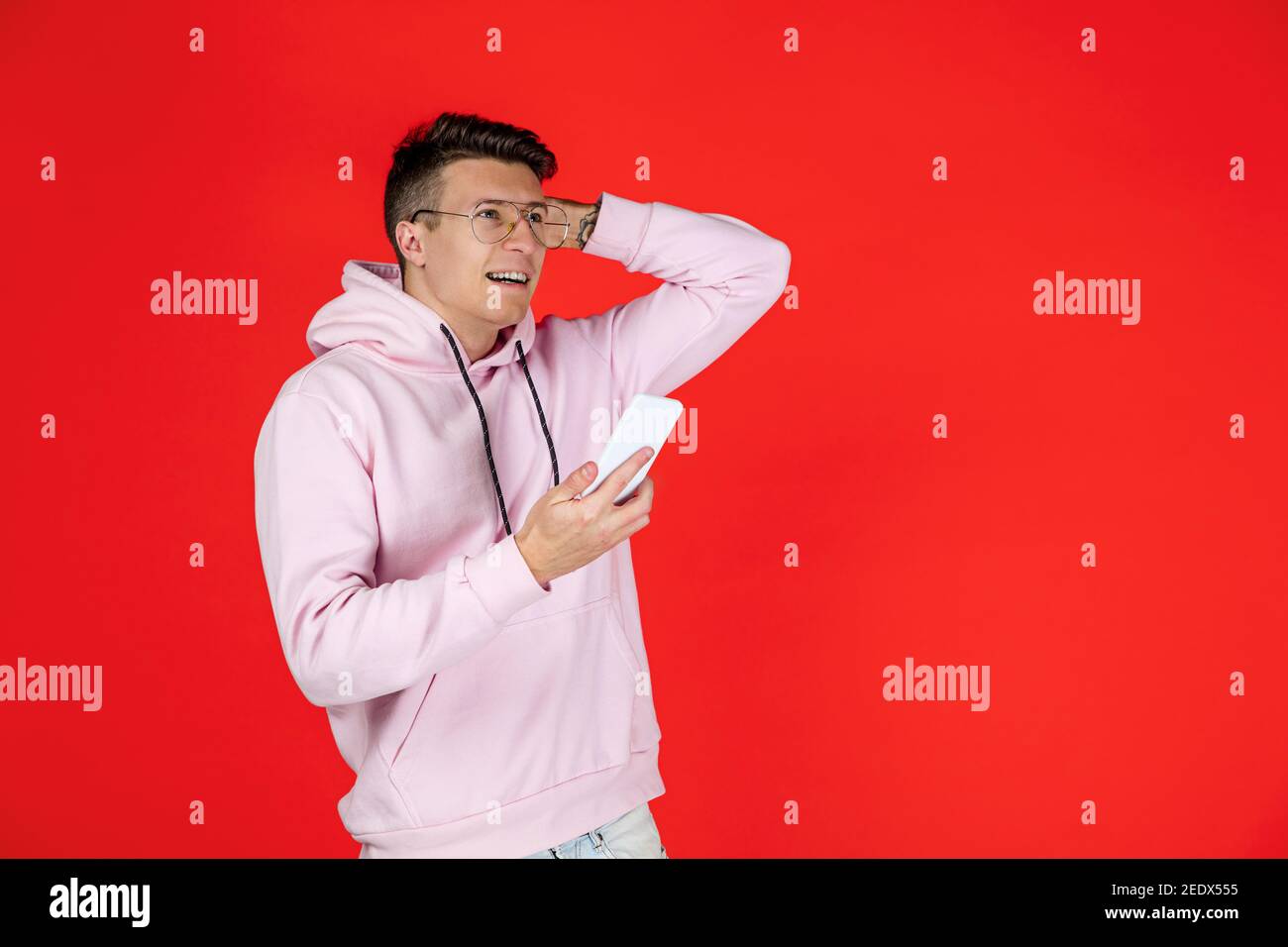 Doubtfully holding phone. Caucasian man's portrait isolated on red background with copyspace. Handsome male model in street style. Concept of human emotions, facial expression, sales, ad, fashion. Stock Photo