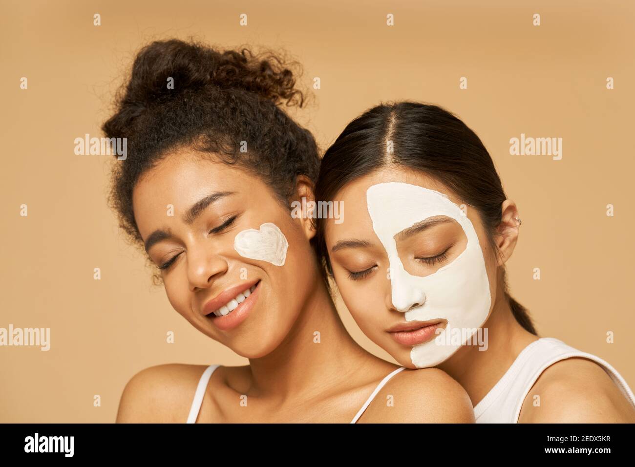 Close up portrait of two young female friends with facial masks on posing with eyes closed isolated over beige background. Skincare, beauty concept Stock Photo