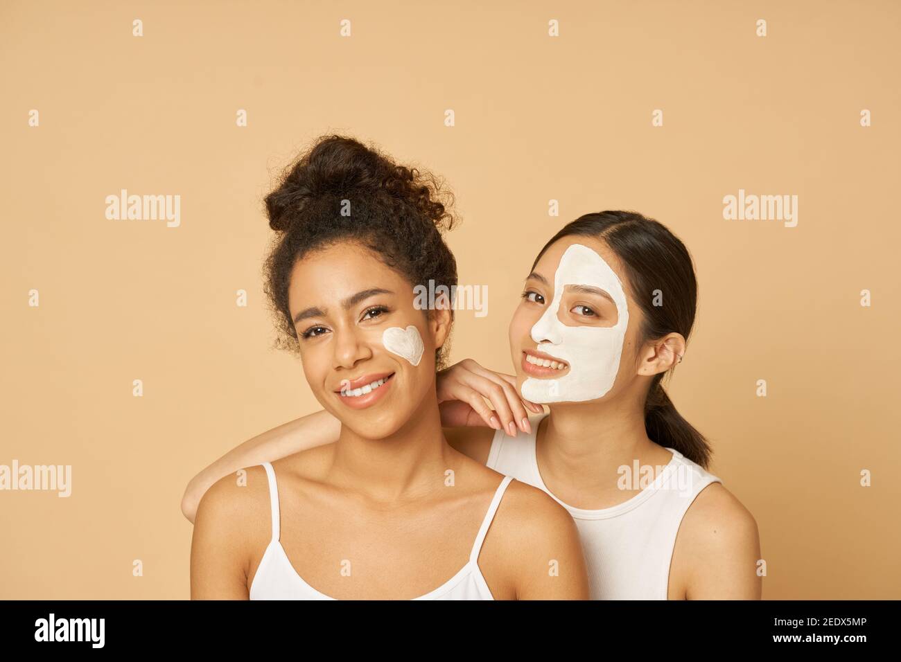 Studio shot of two young women, female friends having fun, smiling at camera while posing with facial masks on isolated over beige background. Horizontal shot Stock Photo