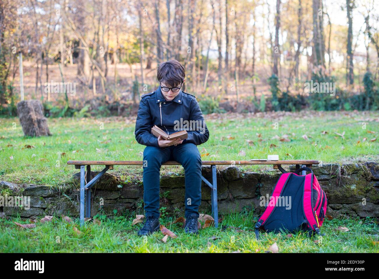 12 year old kid sitting outdoors reads a book, school backpack in the ground-young reader Stock Photo