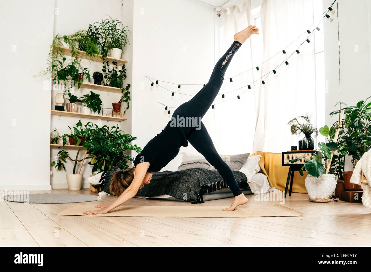 Portrait of beautiful young woman practicing yoga indoor. Woman in black sportswear practice at light home. Bed and green home plants on background. Stock Photo