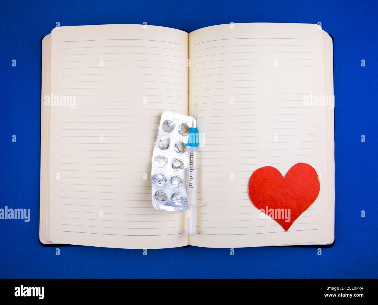 Blank Writing Pad with Heart Shape and Empty Pack of the Pills and a Syringe on the Blue Paper Background closeup Stock Photo