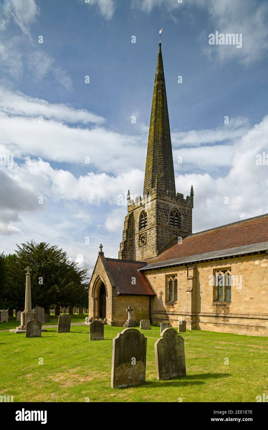Exterior of historic St. Edith's Church (churchyard memorials, porch, tall spire & blue sky) - Bishop Wilton, East Riding of Yorkshire, England, UK. Stock Photo
