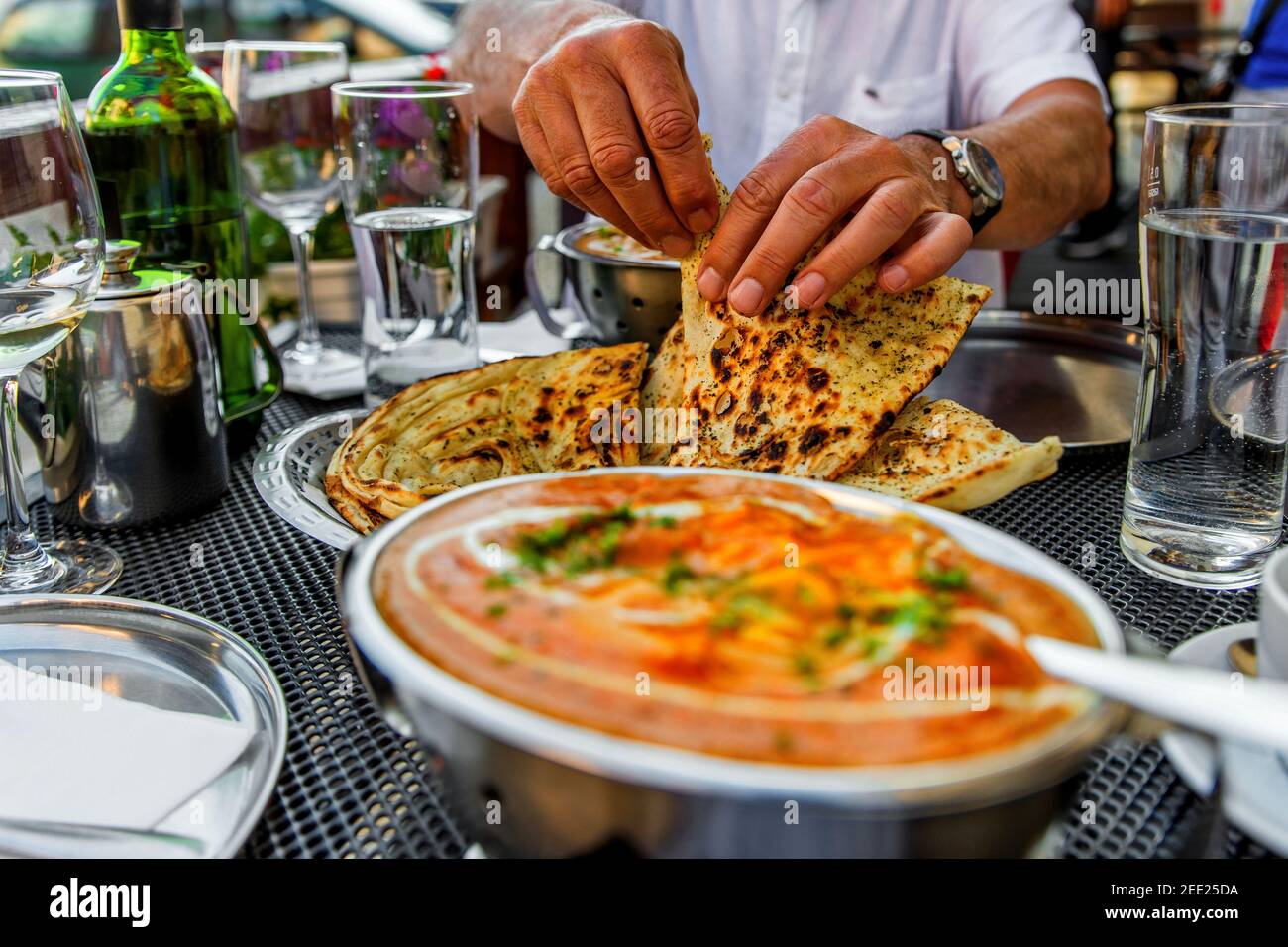 Hands of man tearing bread (butter naan) at the table with indian meals, glasses of water and wine, jug with tea, outdoor in indian restaurant. Stock Photo