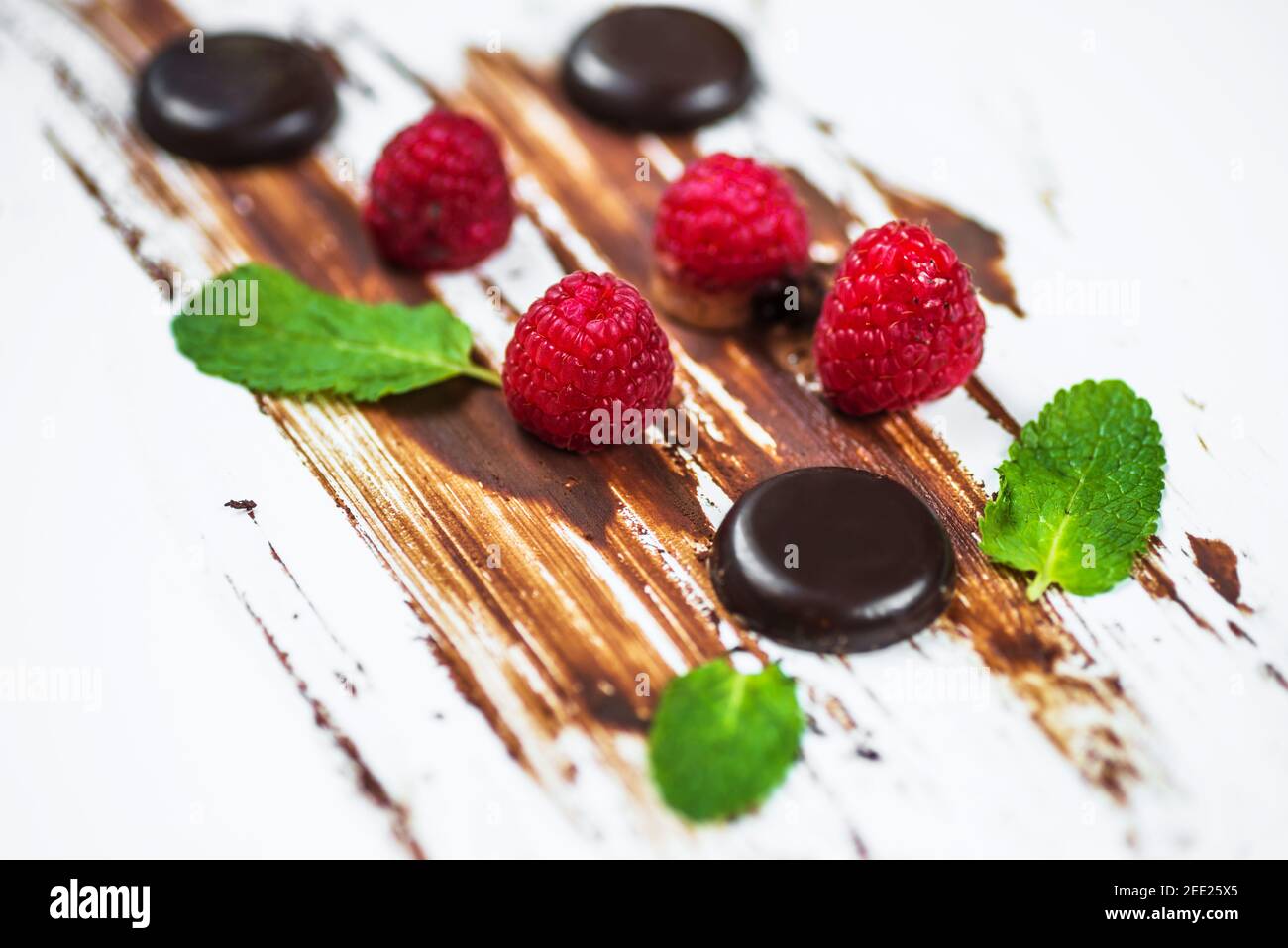 Raspberries, mint leaf and small chocolate wheel (pastille) on chocolate lines on slanting white background. Stock Photo