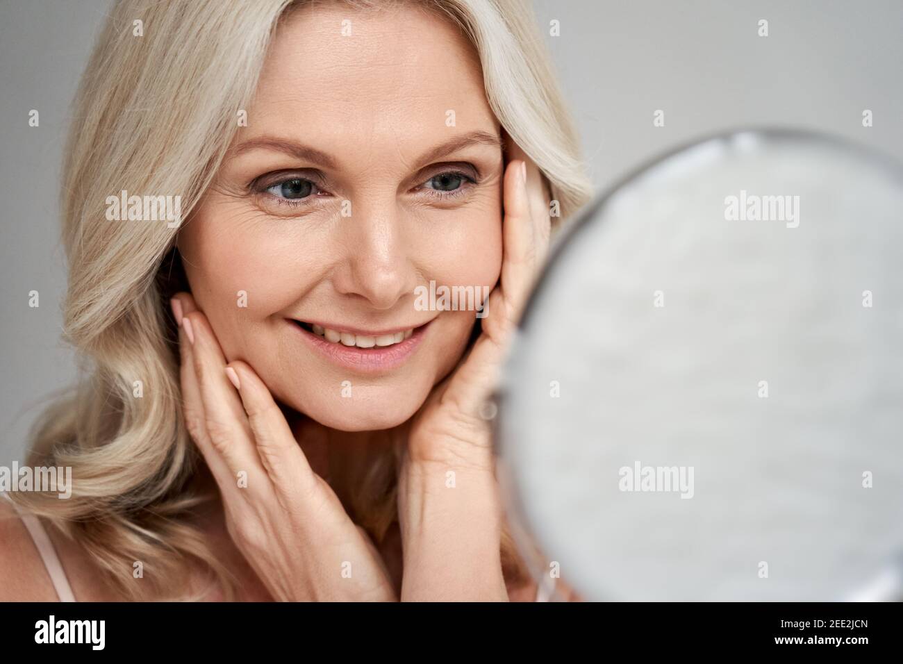 Happy 50s middle aged woman touching face skin looking in mirror. Stock Photo