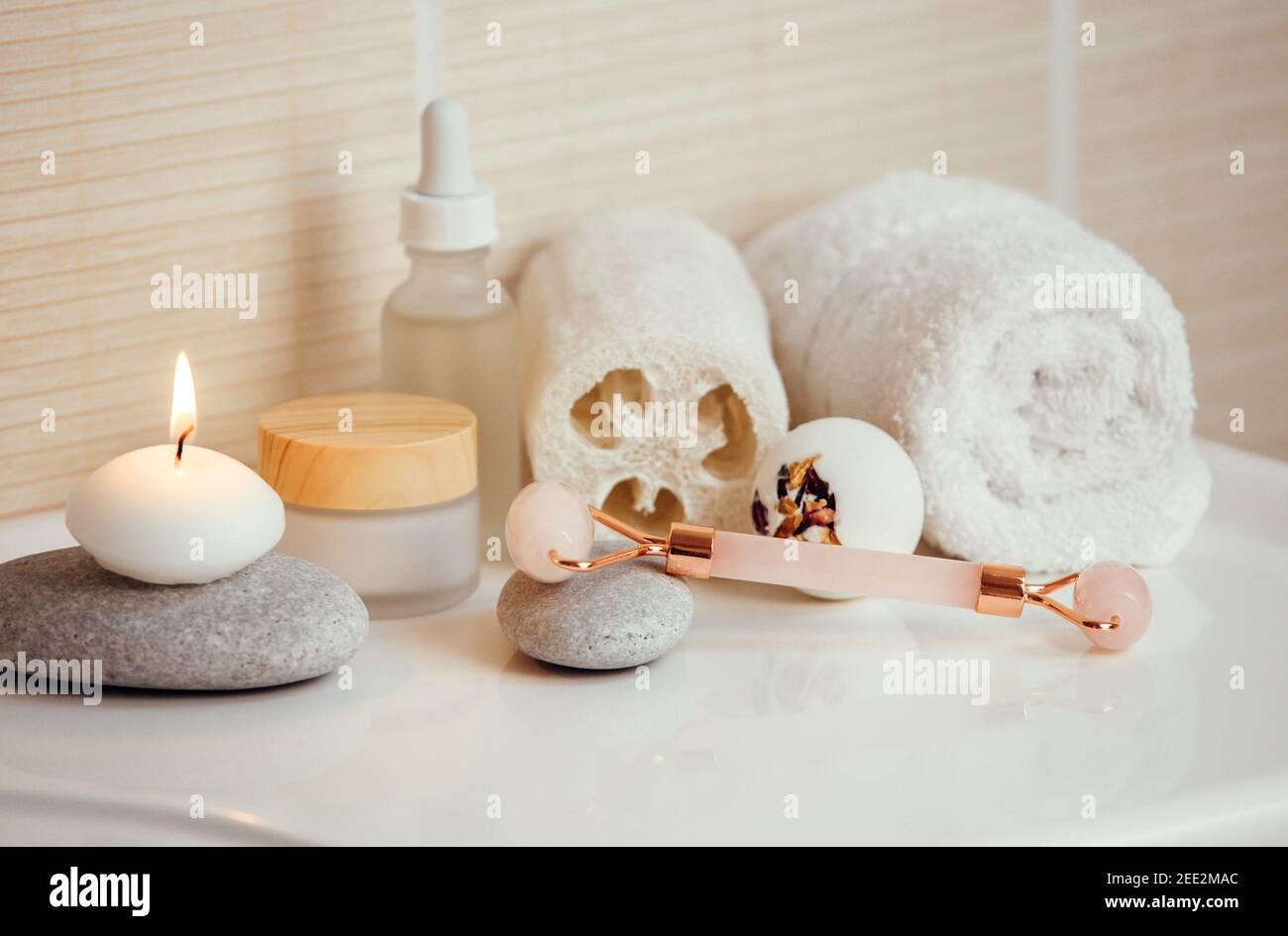 Home spa self care products in bathroom, facial massage rolling tool, moisturizing creams and oil, creamy bath bomb with dry rose petals. Stock Photo