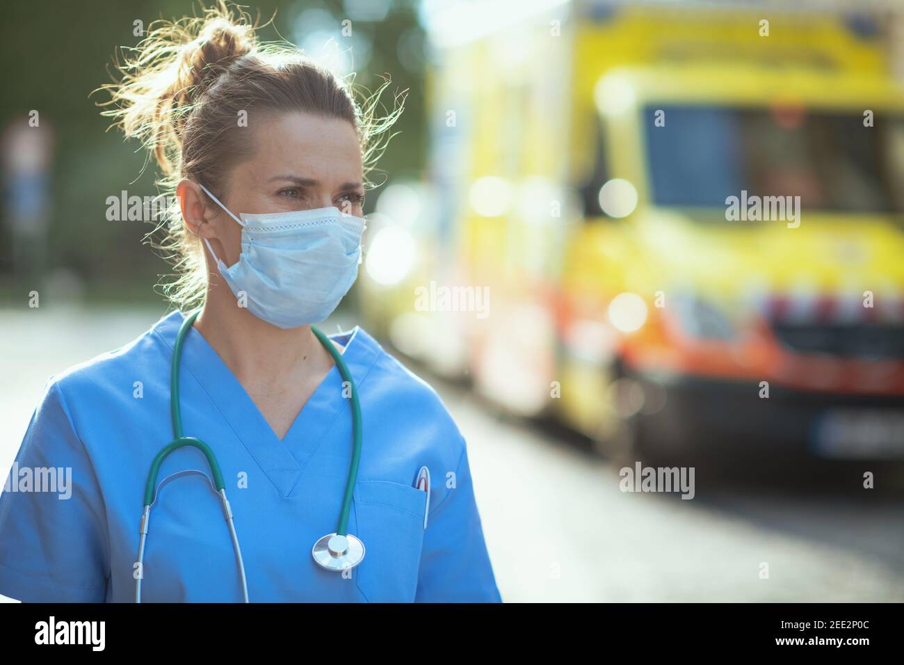 covid-19 pandemic. pensive modern medical doctor woman in uniform with stethoscope and medical mask looking into the distance outside near ambulance. Stock Photo
