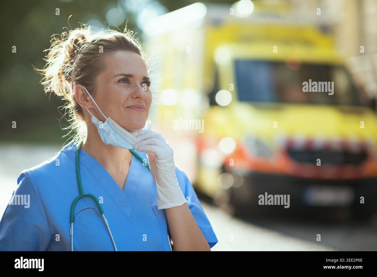 covid-19 pandemic. smiling modern medical doctor woman in scrubs with stethoscope and medical mask breathing outdoors near ambulance. Stock Photo