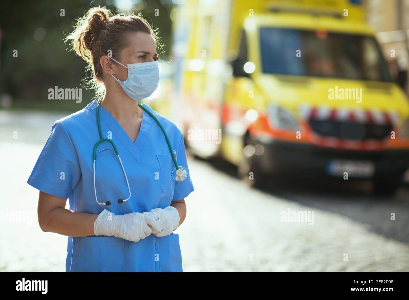 covid-19 pandemic. pensive modern medical doctor woman in uniform with stethoscope and medical mask outdoors near ambulance. Stock Photo