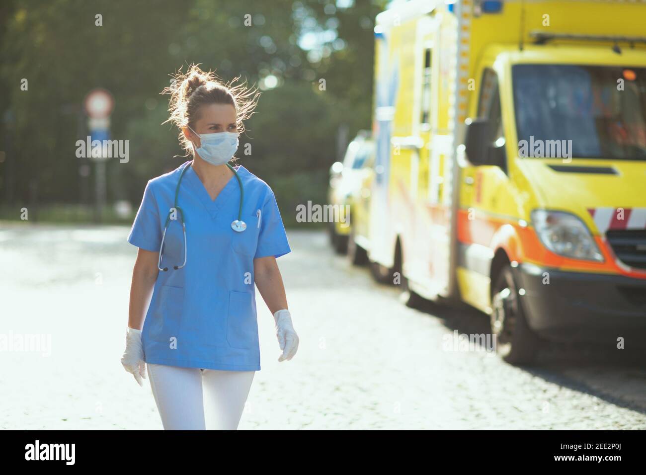 coronavirus pandemic. modern paramedic woman in scrubs with stethoscope and medical mask walking outside near ambulance. Stock Photo