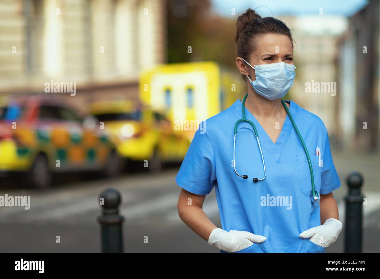covid-19 pandemic. modern medical doctor woman in uniform with stethoscope and medical mask looking into the distance outside near ambulance. Stock Photo
