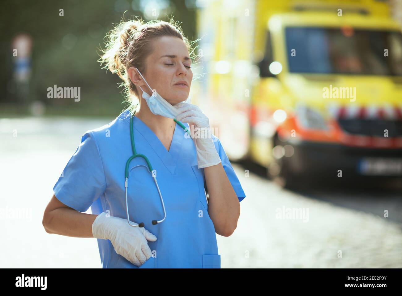 coronavirus pandemic. tired modern paramedic woman in scrubs with stethoscope and medical mask breathing outside near ambulance. Stock Photo