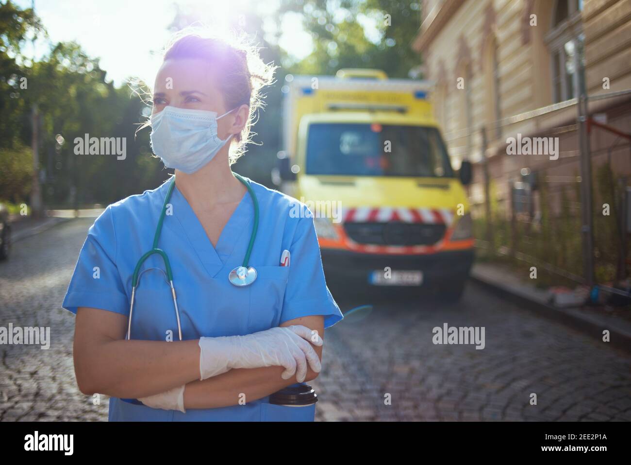 covid-19 pandemic. modern medical doctor woman in uniform with stethoscope and medical mask looking into the distance outdoors near ambulance. Stock Photo