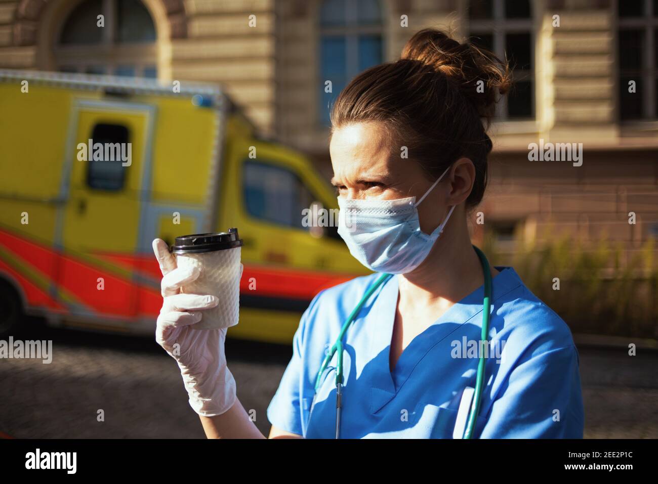 covid-19 pandemic. tired modern medical doctor woman in uniform with stethoscope, medical mask and cup of coffee outside near ambulance. Stock Photo