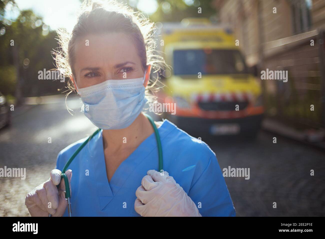covid-19 pandemic. pensive modern paramedic woman in uniform with stethoscope and medical mask outside near ambulance. Stock Photo