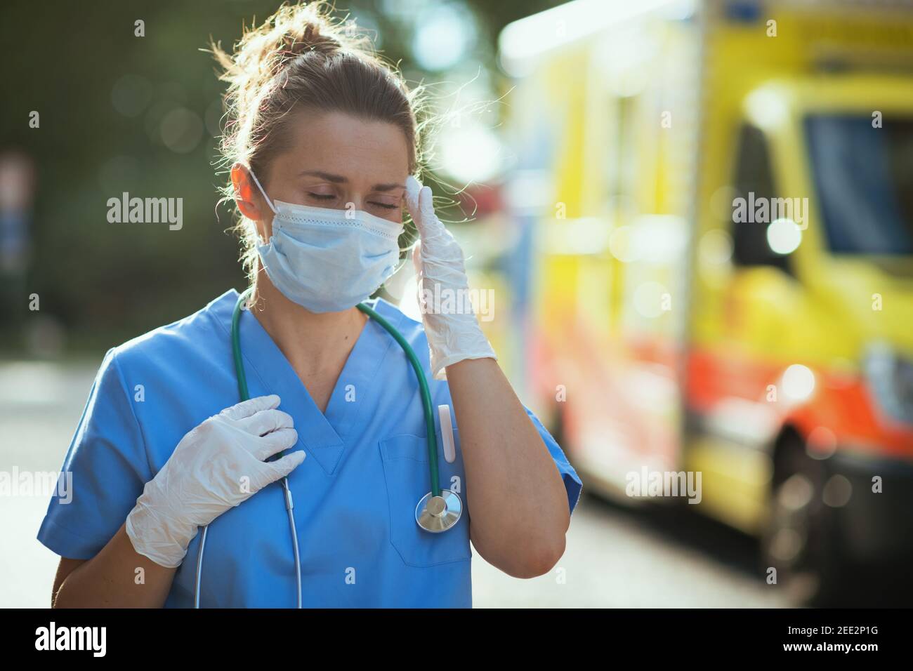 coronavirus pandemic. stressed modern paramedic woman in scrubs with stethoscope and medical mask outdoors near ambulance. Stock Photo