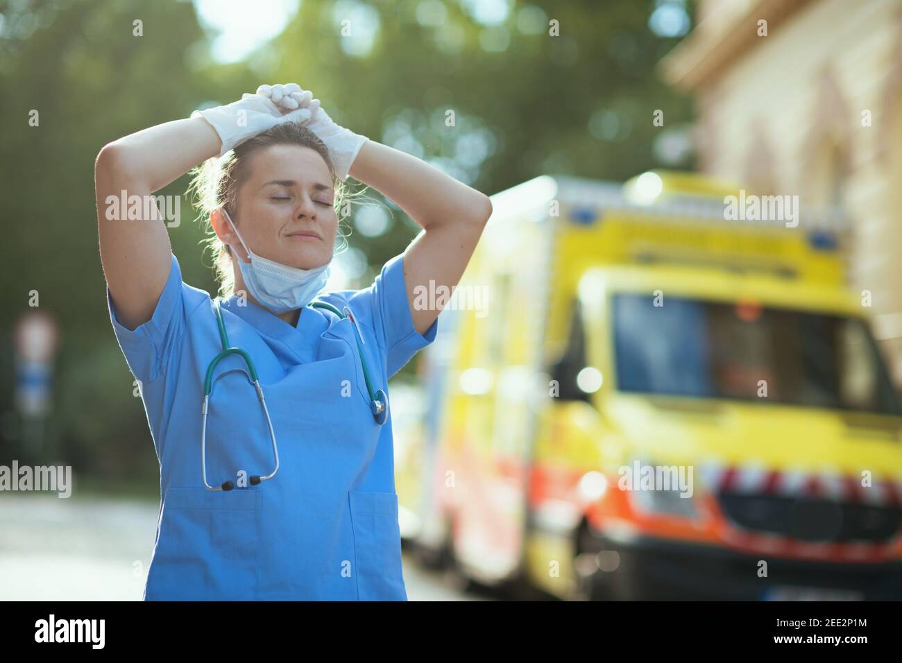 coronavirus pandemic. relaxed modern paramedic woman in uniform with stethoscope and medical mask breathing outdoors near ambulance. Stock Photo