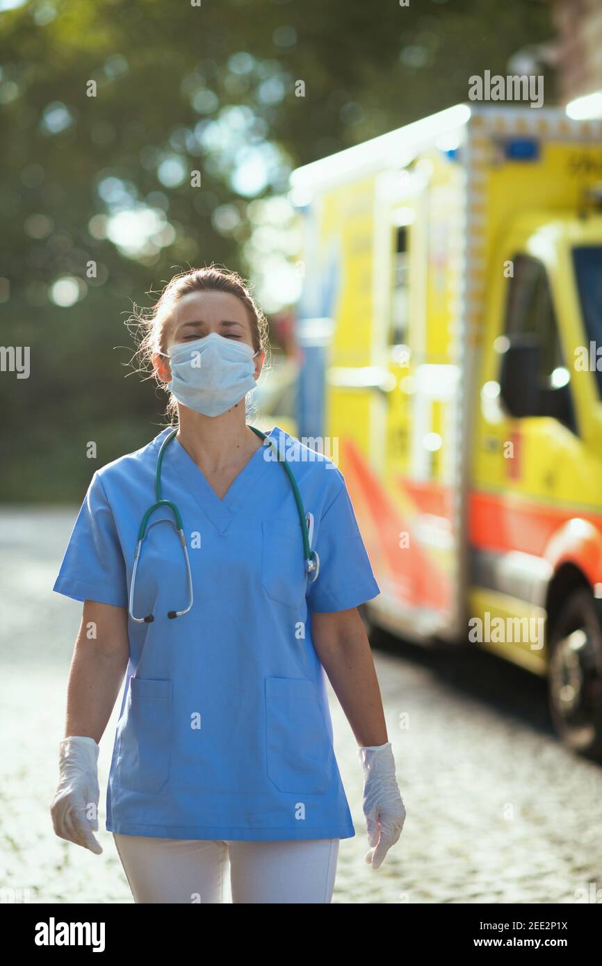 coronavirus pandemic. modern medical doctor woman in scrubs with stethoscope and medical mask outdoors near ambulance. Stock Photo