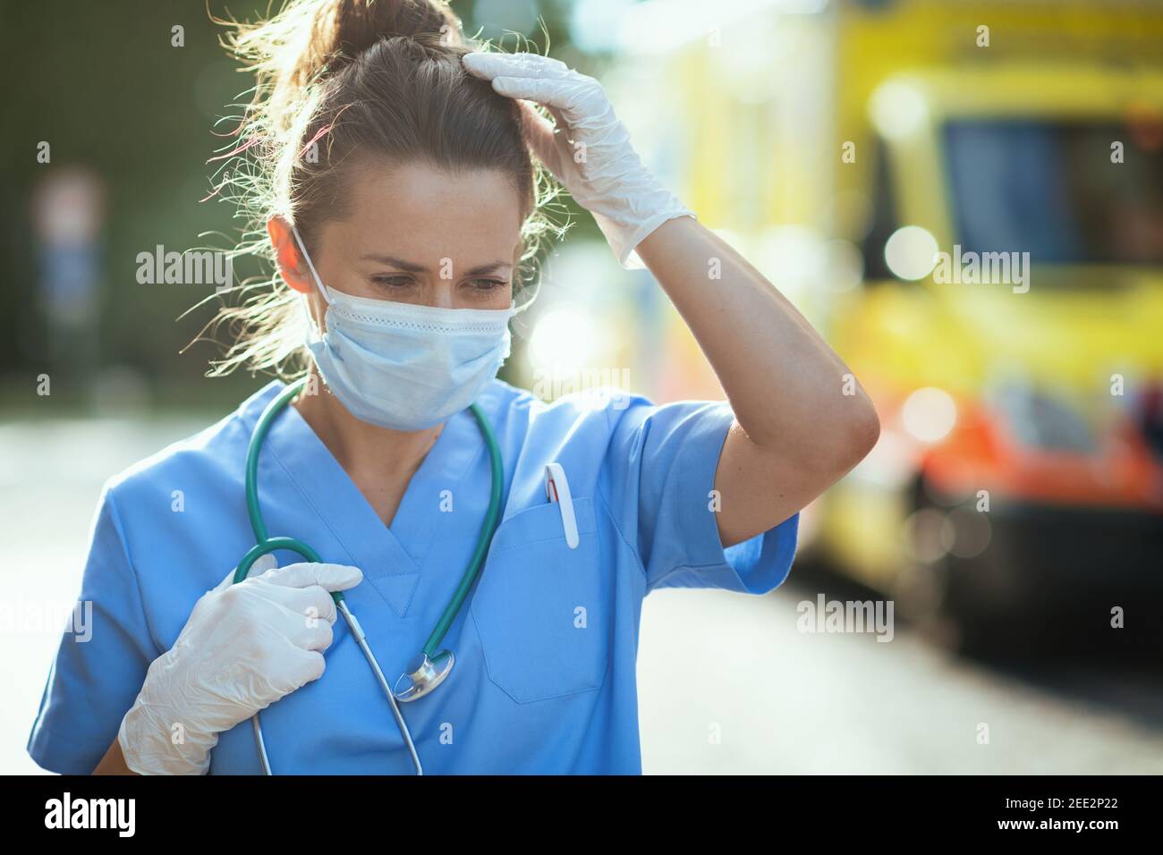 coronavirus pandemic. unhappy modern paramedic woman in scrubs with stethoscope and medical mask outside near ambulance. Stock Photo