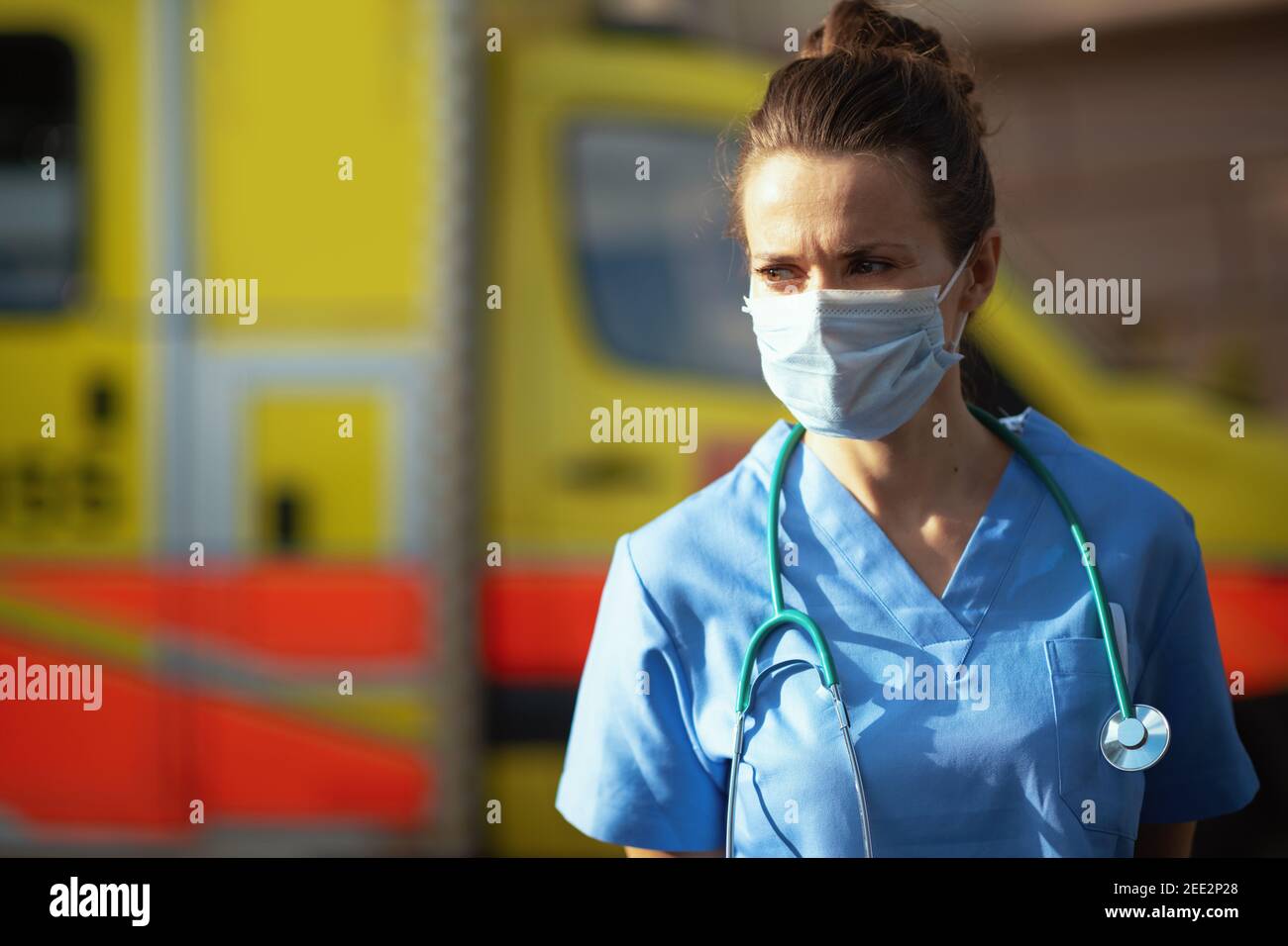 covid-19 pandemic. pensive modern paramedic woman in uniform with stethoscope and medical mask outdoors near ambulance. Stock Photo