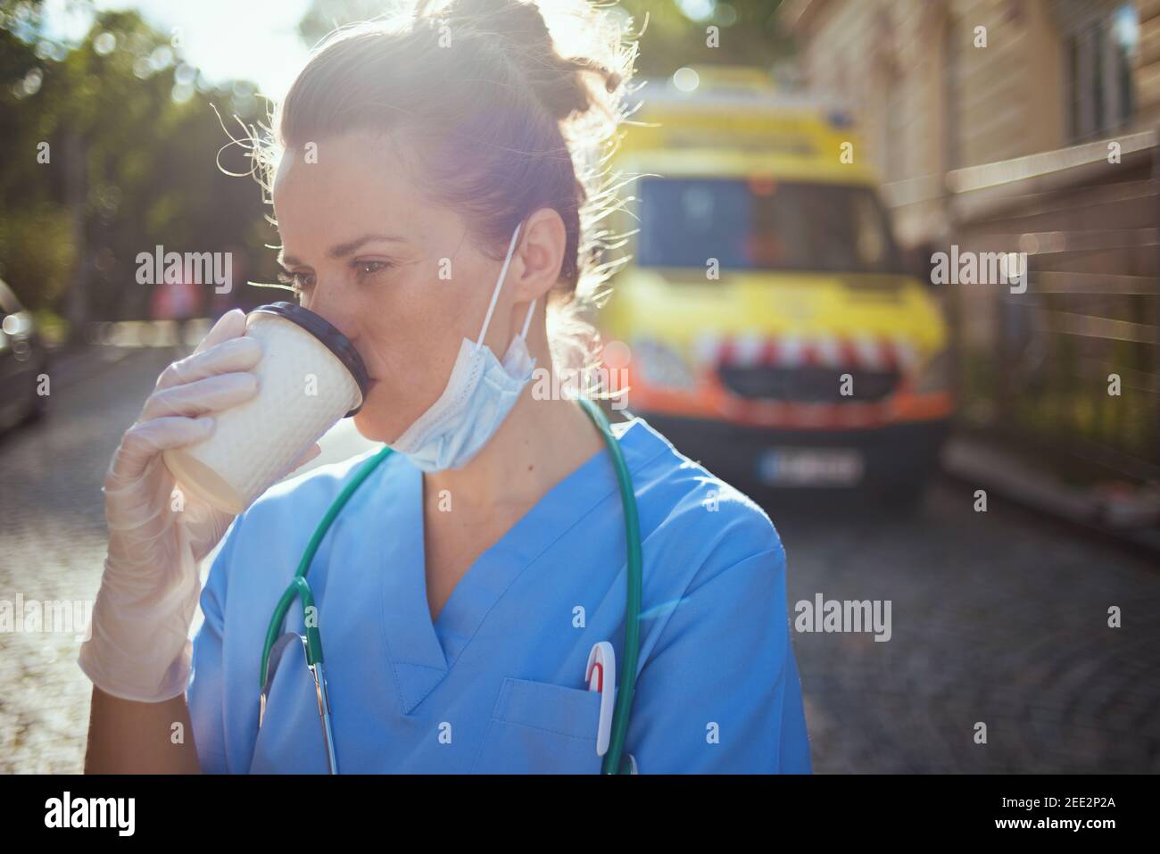 covid-19 pandemic. tired modern medical doctor woman in uniform with stethoscope, medical mask and cup of coffee outside near ambulance. Stock Photo