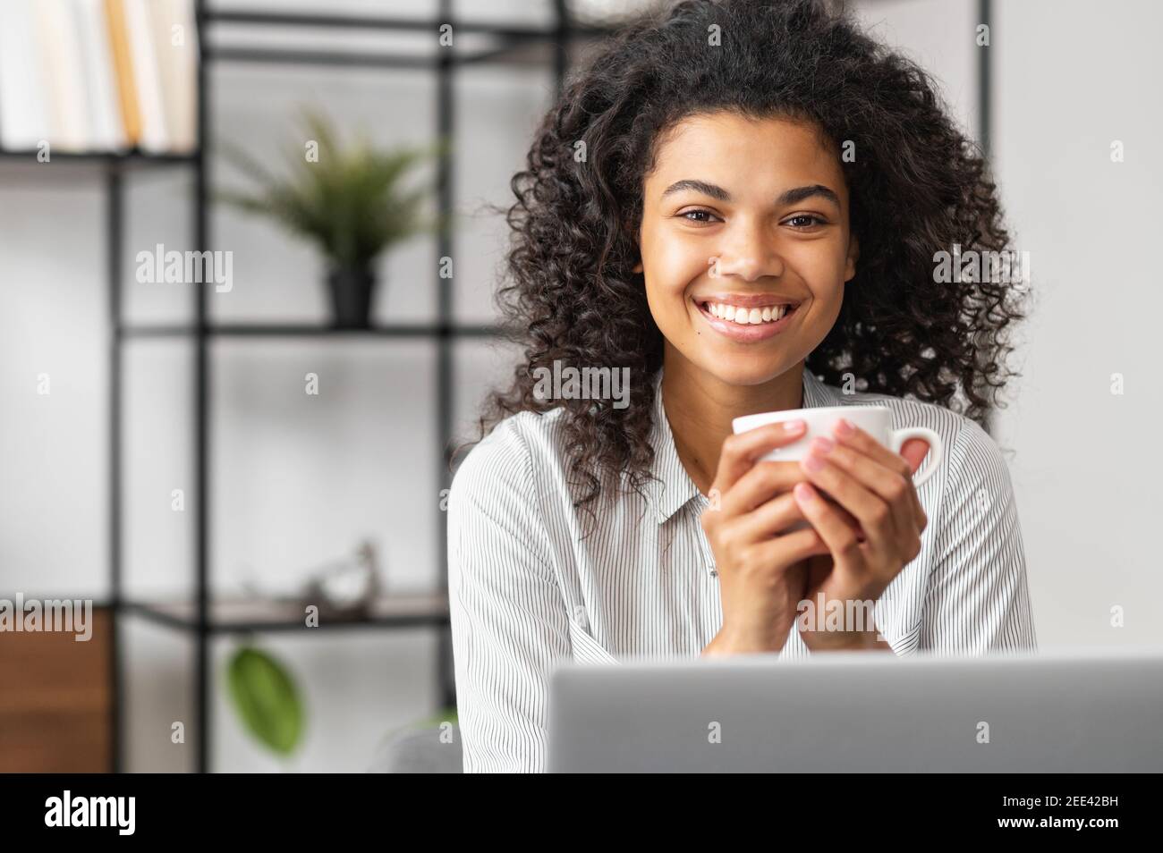 Smiling young cheerful African American woman with Afro hairstyle enjoying morning coffee while working on the laptop, sitting at the desk in the home Stock Photo