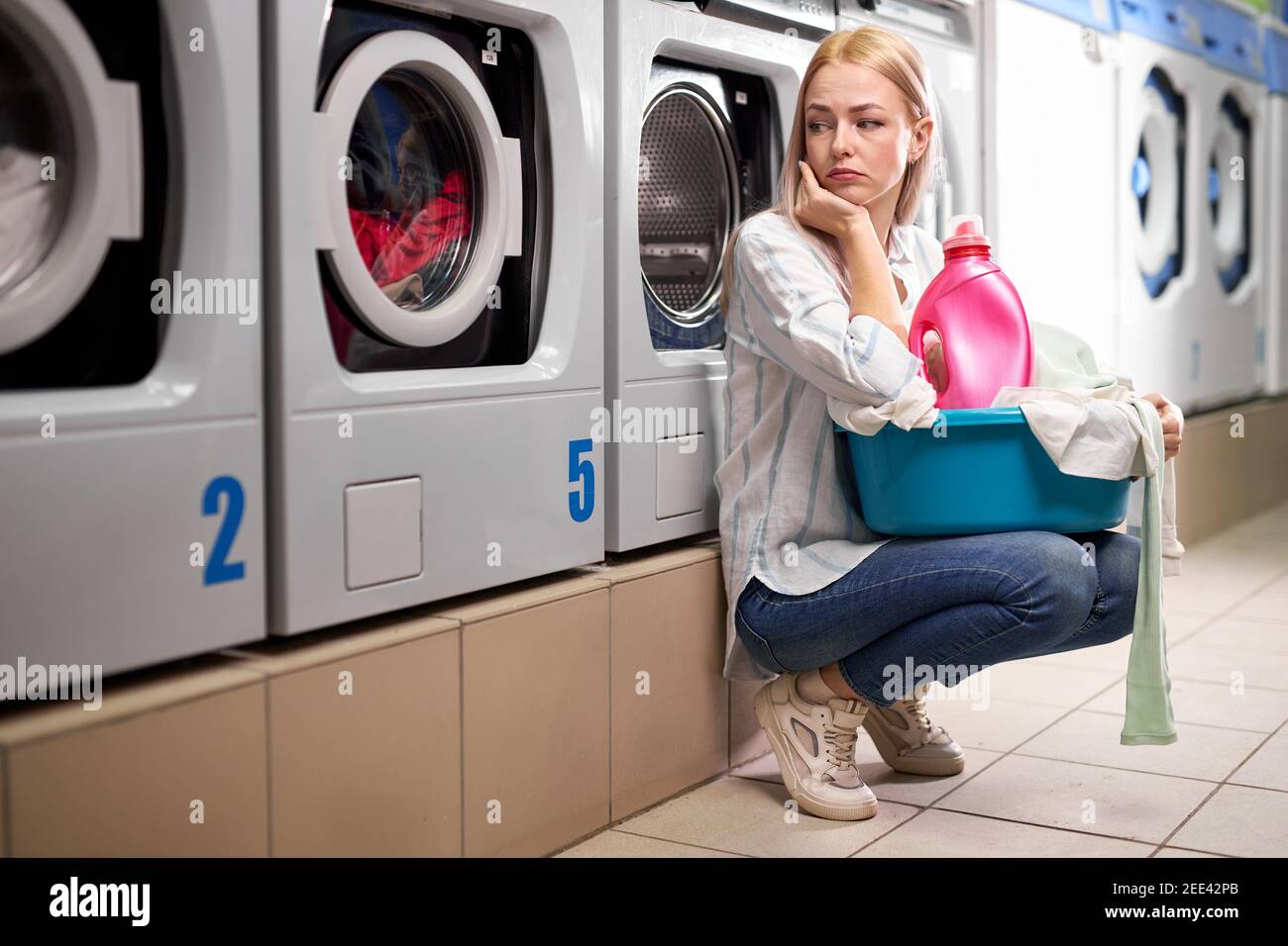 tired woman sitting near washing machines and looking up sadly. Caucasian woman spent time on washing, waiting and bored female with dirty clothes in Stock Photo
