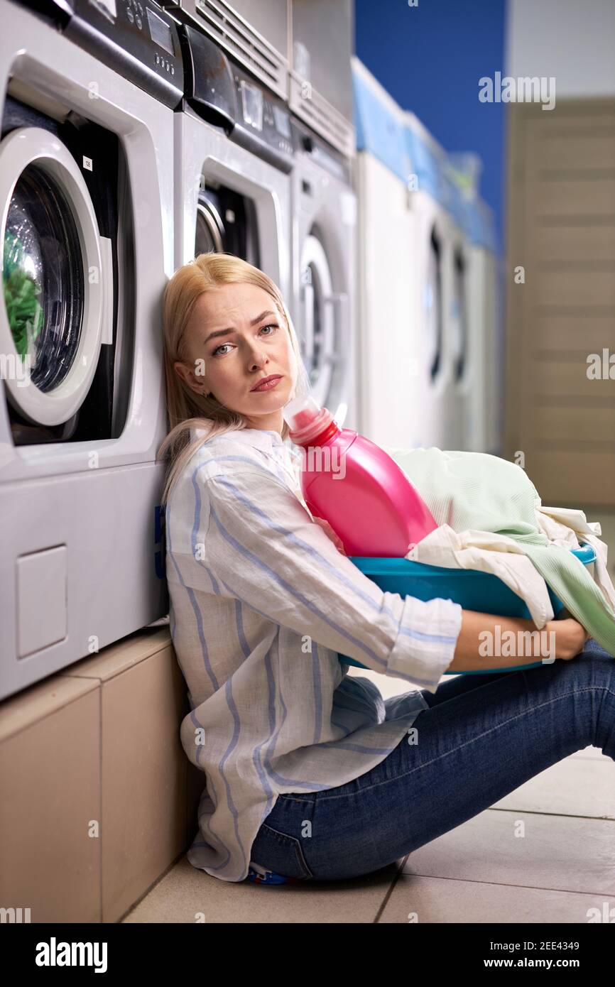 Bored Woman Sitting By Washing Machine, Waiting For End of Washing, Blond Caucasian Lady holding basin or basket with remaining clothes for washing an Stock Photo