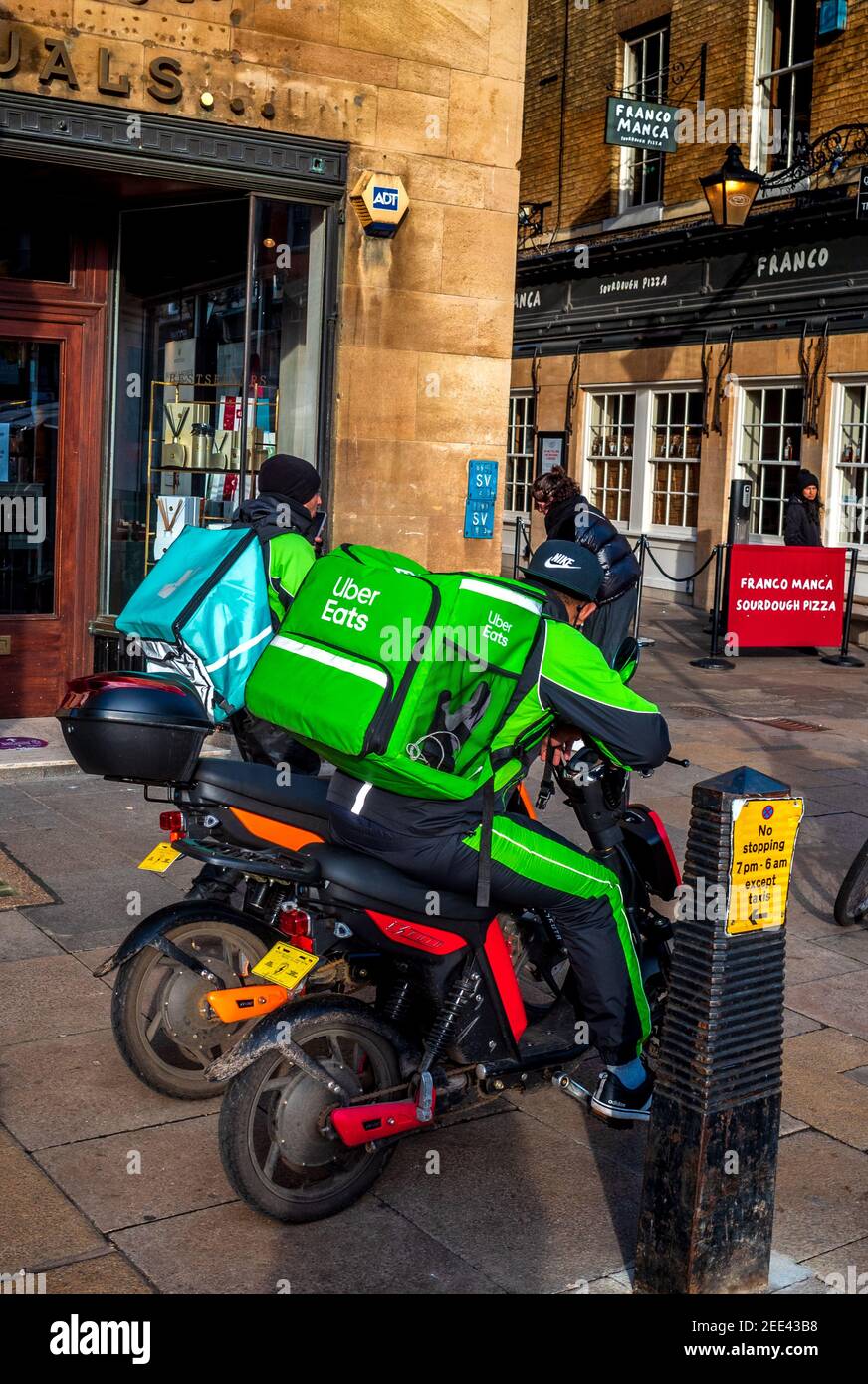UBER Eats and Deliveroo food delivery couriers wait for order pickup outide the Franco Manca Pizzeria in Cambridge UK. Stock Photo