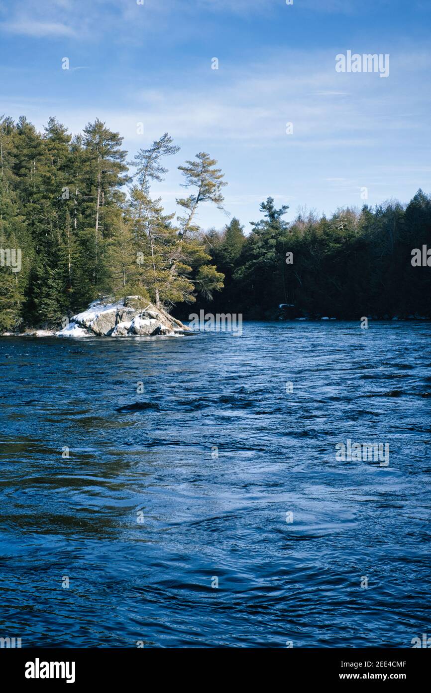 Near Burnstown, Ontario, rapids along the Madawaska River flow around a rocky island, dusted with snow and supporting evergreen trees. Stock Photo