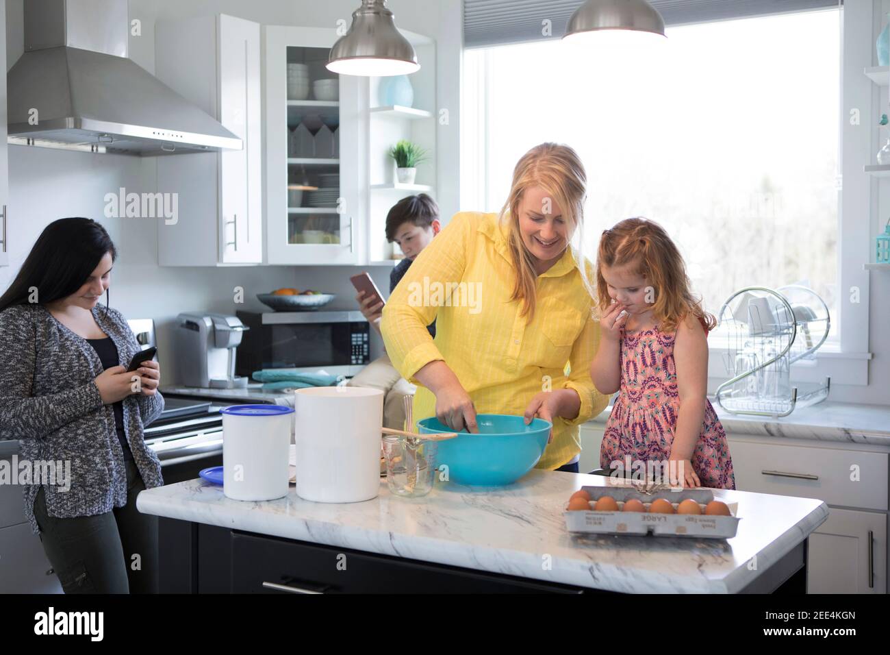 A mom and toddler daughter bake together while the teenage siblings text on their phones. Stock Photo