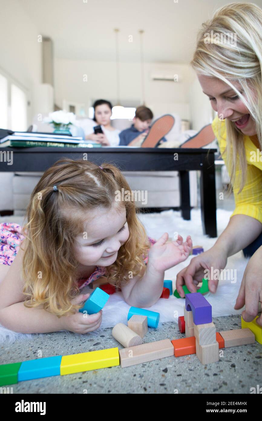 A mom and toddler daughter play with toys on the floor of a modern home. Teen siblings look at phones in the background. Stock Photo