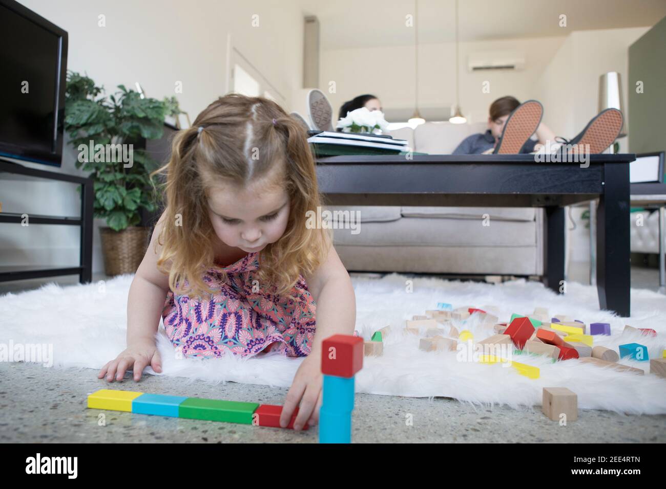 Little girl playing with blocks on the floor. Teenage siblings sit on the couch behind her. Stock Photo