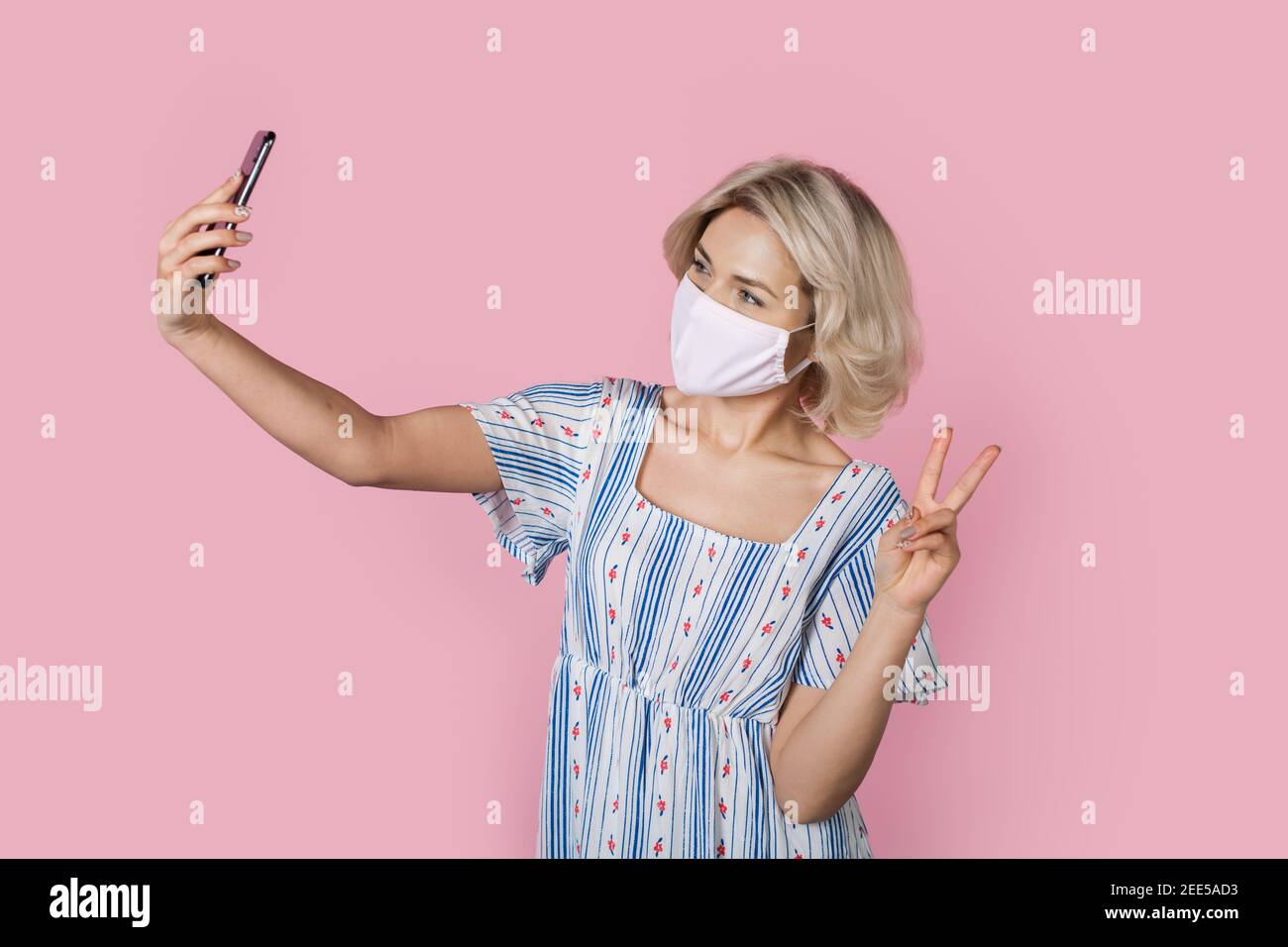 Adorable blonde lady in medical mask in gesturing the peace sign while taking a selfie on a pink wall Stock Photo