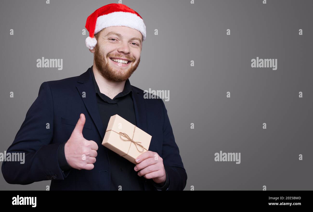 Ginger man with beard and santa hat is gesturing the like sign on a gray wall holding a present and smile Stock Photo
