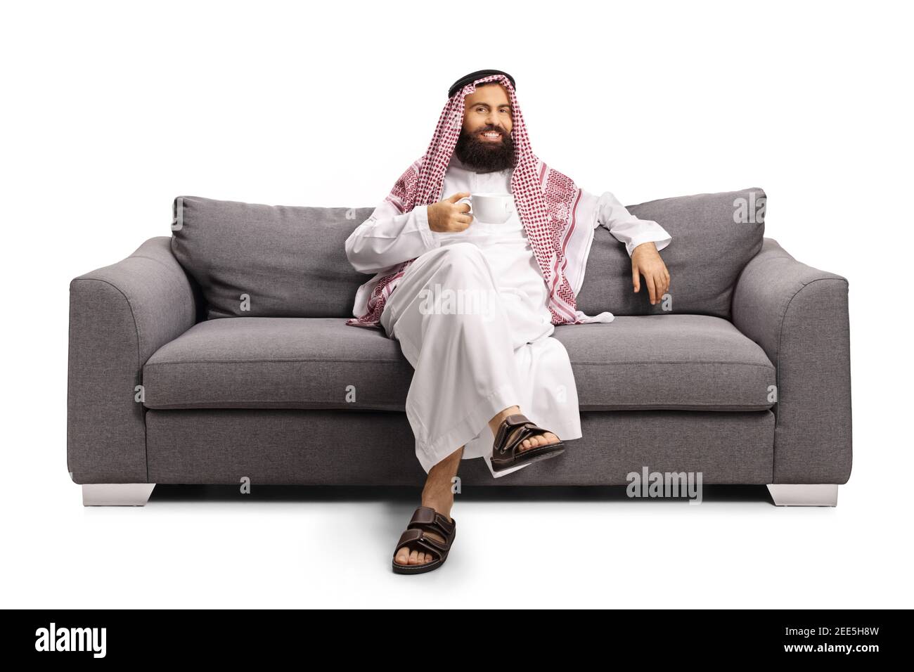 Saudi arab man sitting on a gray sofa and drinking a cup of tea isolated on white background Stock Photo