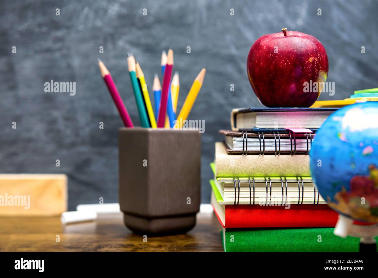 Stack of colorful books, stationery and education supplies on wooden desk in classroom with blackboard in background Stock Photo