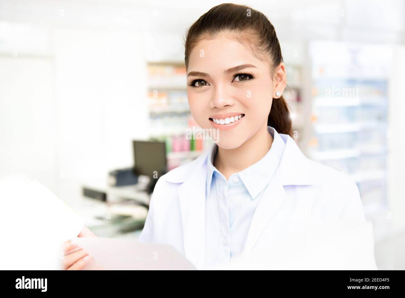 Smiling young Asian woman pharmacist in white gown coat looking at camera while working in pharmacy (chemist shop or drugstore) Stock Photo