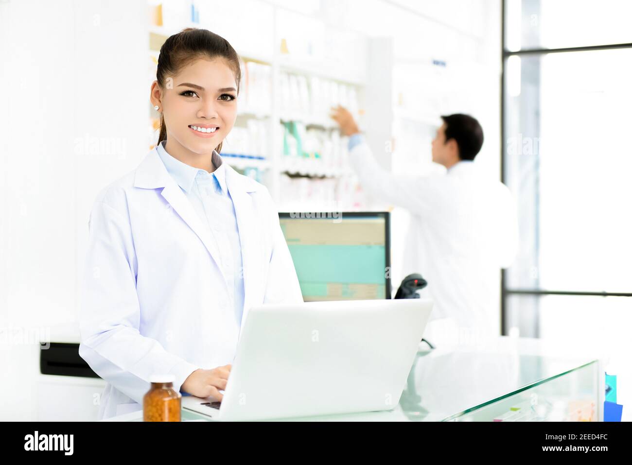 Young smiling Asian woman pharmacist in white gown coat uniform working on laptop computer in chemist shop or pharmacy Stock Photo