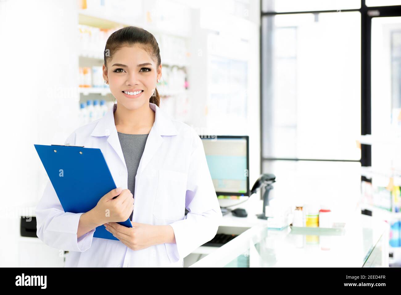 Smiling beautiful young Asian woman pharmacist in white gown coat at working in pharmacy (chemist shop or drugstore) Stock Photo