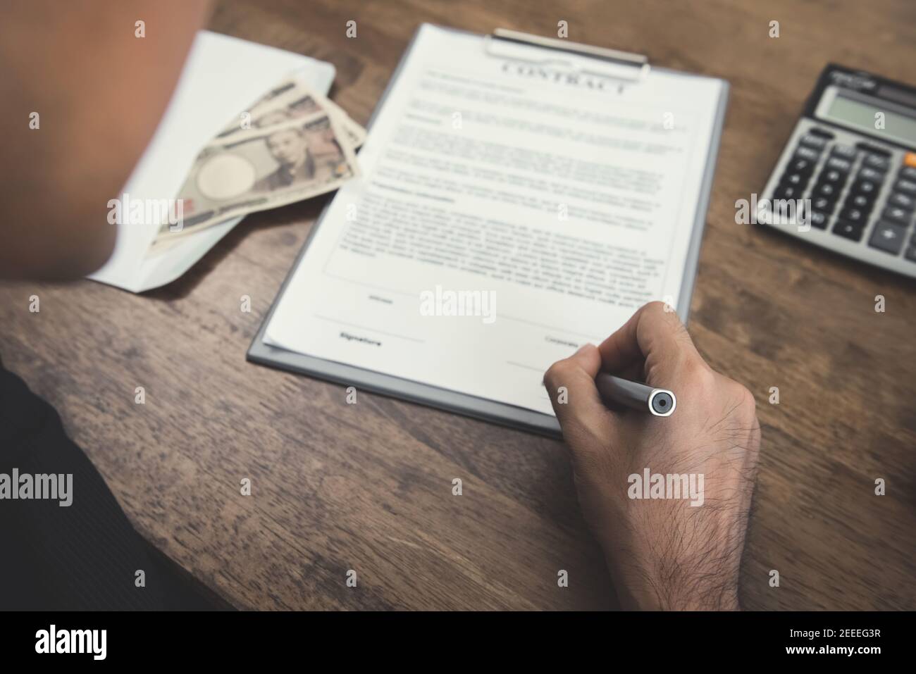 Hand holding pen signing contract with money, Japanese yen banknotes, on the table Stock Photo