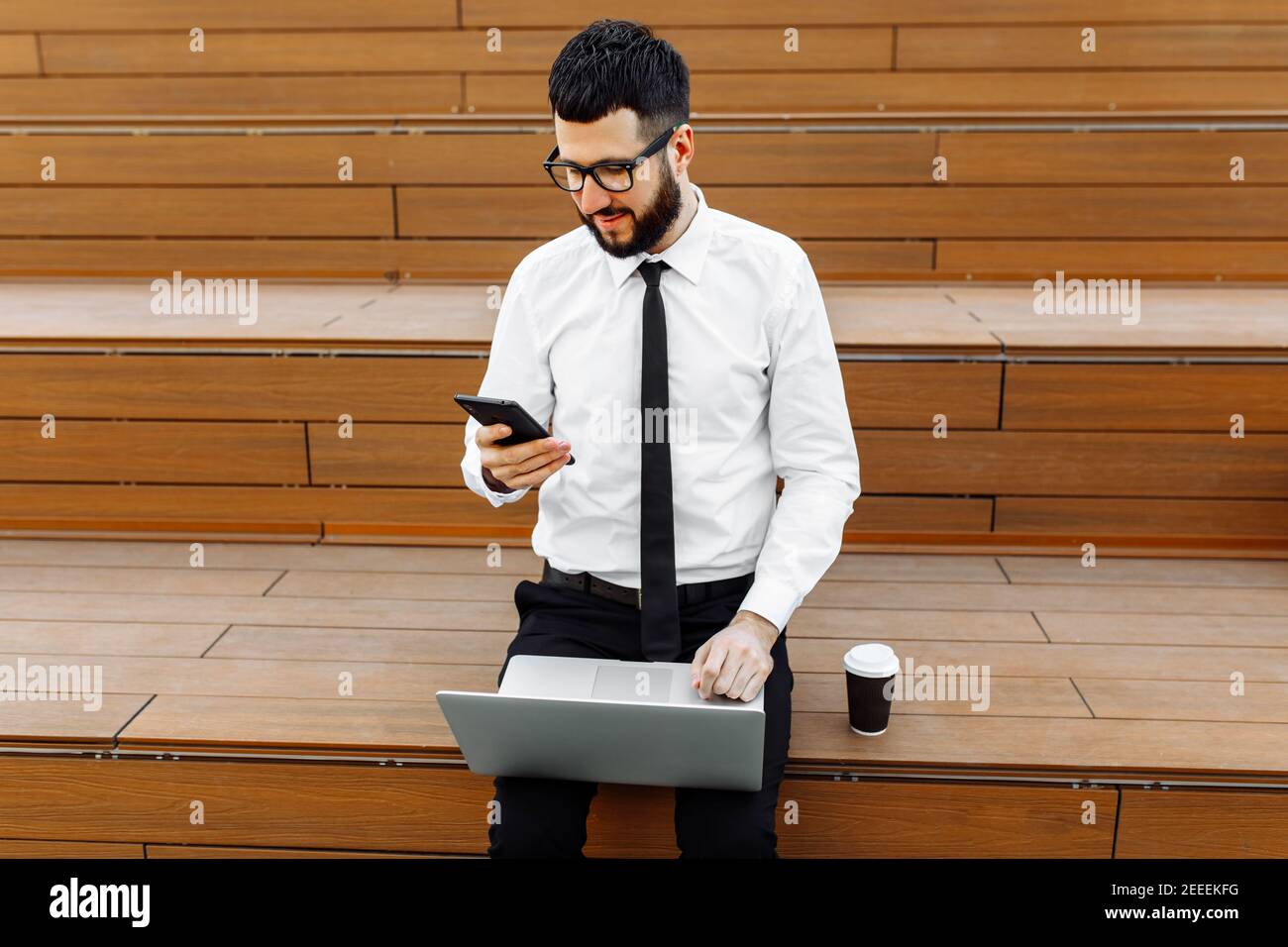 Young handsome businessman in glasses sits on a bench with a laptop and uses a mobile phone outdoors in the city Stock Photo