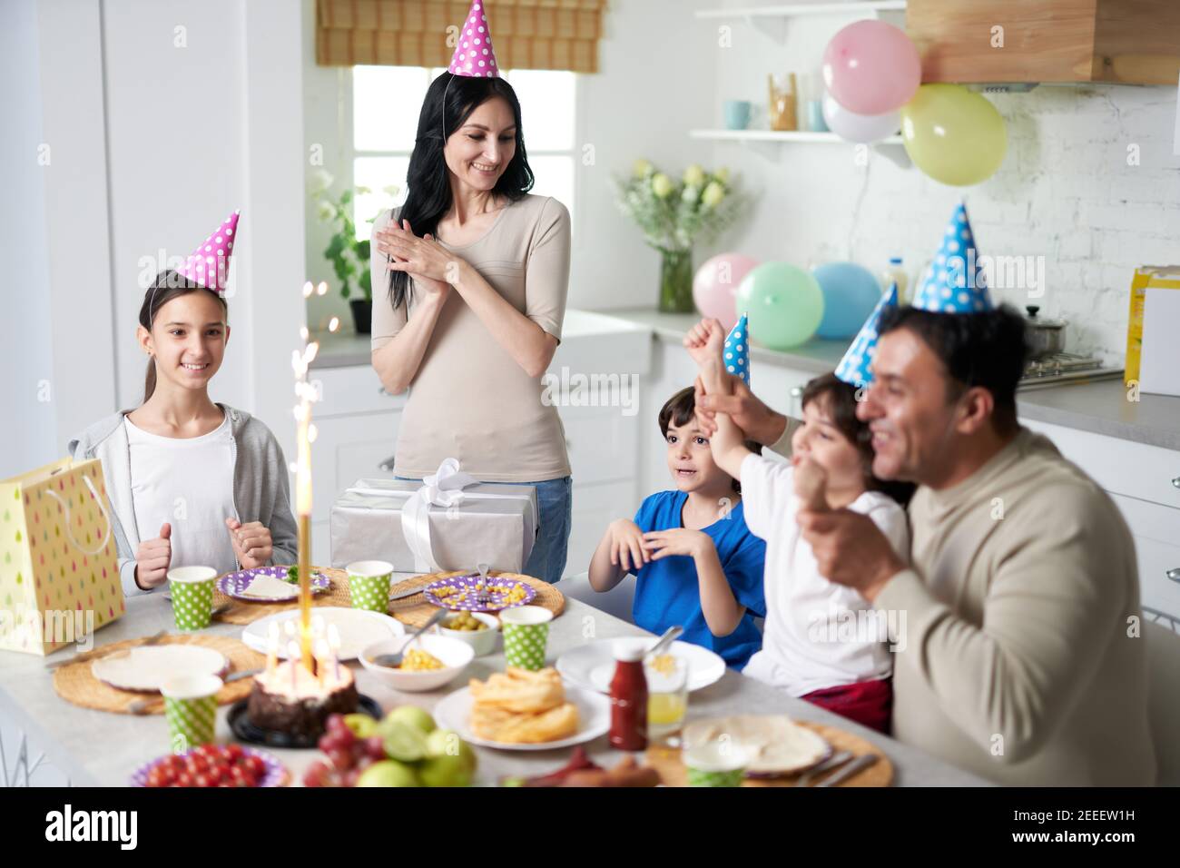Cheerful latin family with children wearing birthday caps on heads looking happy while celebrating birthday together at home. Parenthood, celebration concept Stock Photo