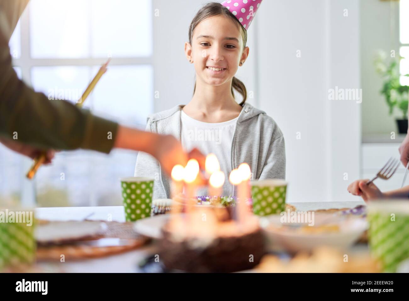 Happy teenage latin girl in birtday cap smiling and looking at birthday cake while celebrating birthday with her family at home. Kids, celebration concept Stock Photo