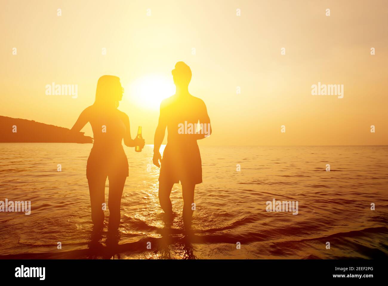 Silhouette of couple holding beer bottles walking in seawater at the beach in twilight sunset Stock Photo